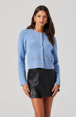 A woman is wearing a light blue textured cardigan with long sleeves and gold buttons down the front. The cardigan has a round neckline and two small pockets at the front. She pairs it with a high-waisted black leather mini skirt with front pleats and side slits.