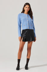 A woman is wearing a light blue, textured, button-up cardigan with long sleeves and gold buttons. She pairs it with a black, high-waisted, leather-like pleated skirt that has a side slit. She completes her outfit with black socks and black loafers, along with gold hoop earrings.