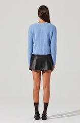 A woman is wearing a light blue, soft and textured long-sleeve sweater with a round neckline. She pairs it with a short, black leather skirt that has a slightly flared silhouette. She completes her outfit with black ankle socks and black loafers.