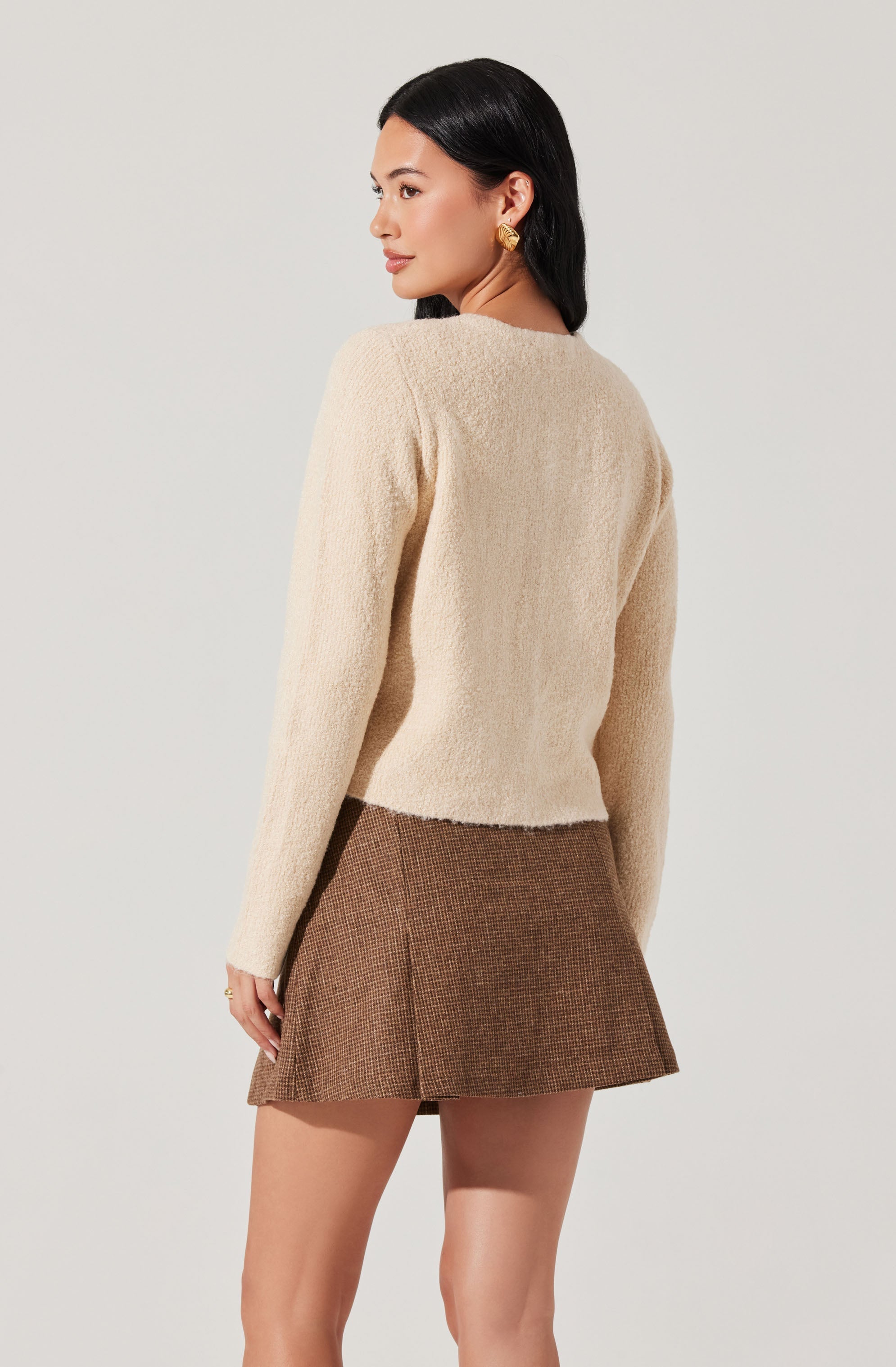 A woman is wearing a beige, textured, long-sleeved sweater and a brown, plaid, A-line mini skirt.