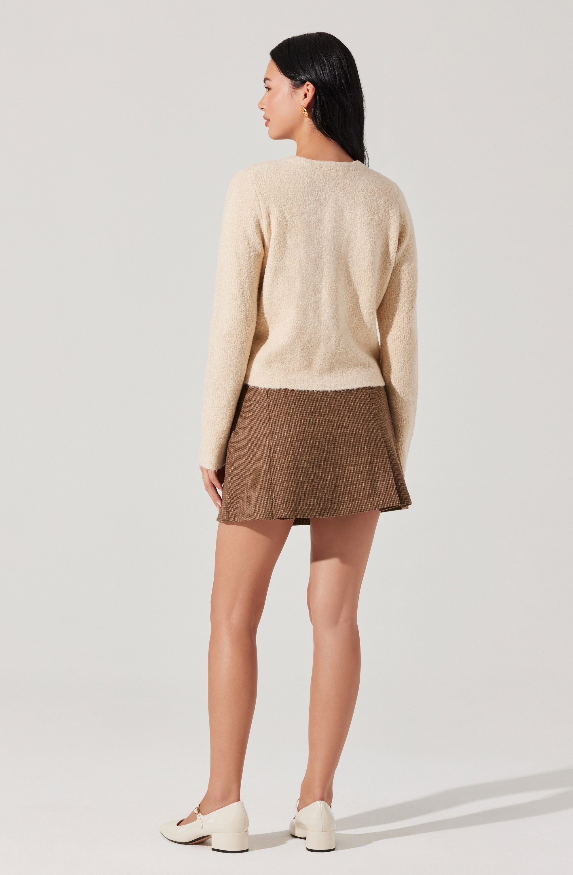 A woman is wearing a long-sleeved, cream-colored, fuzzy sweater paired with a brown, textured, A-line mini skirt. She has on white shoes with a chunky heel and is accessorized with earrings.