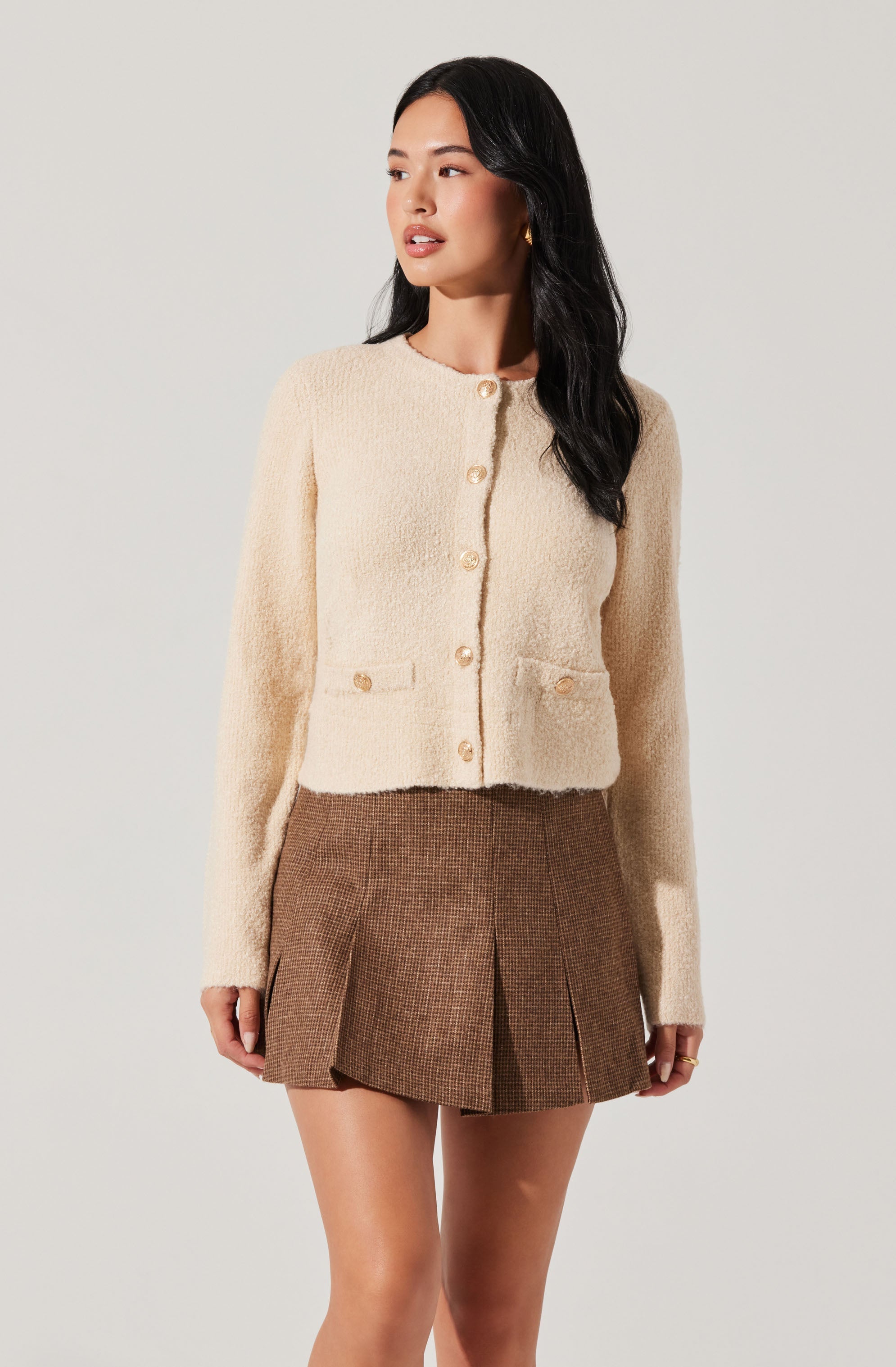 A woman wearing a cream-colored, textured, long-sleeve button-up cardigan with a round neckline and two small decorative pockets at the front. She pairs it with a high-waisted, brown plaid mini skirt that features pleats and falls above the knees.