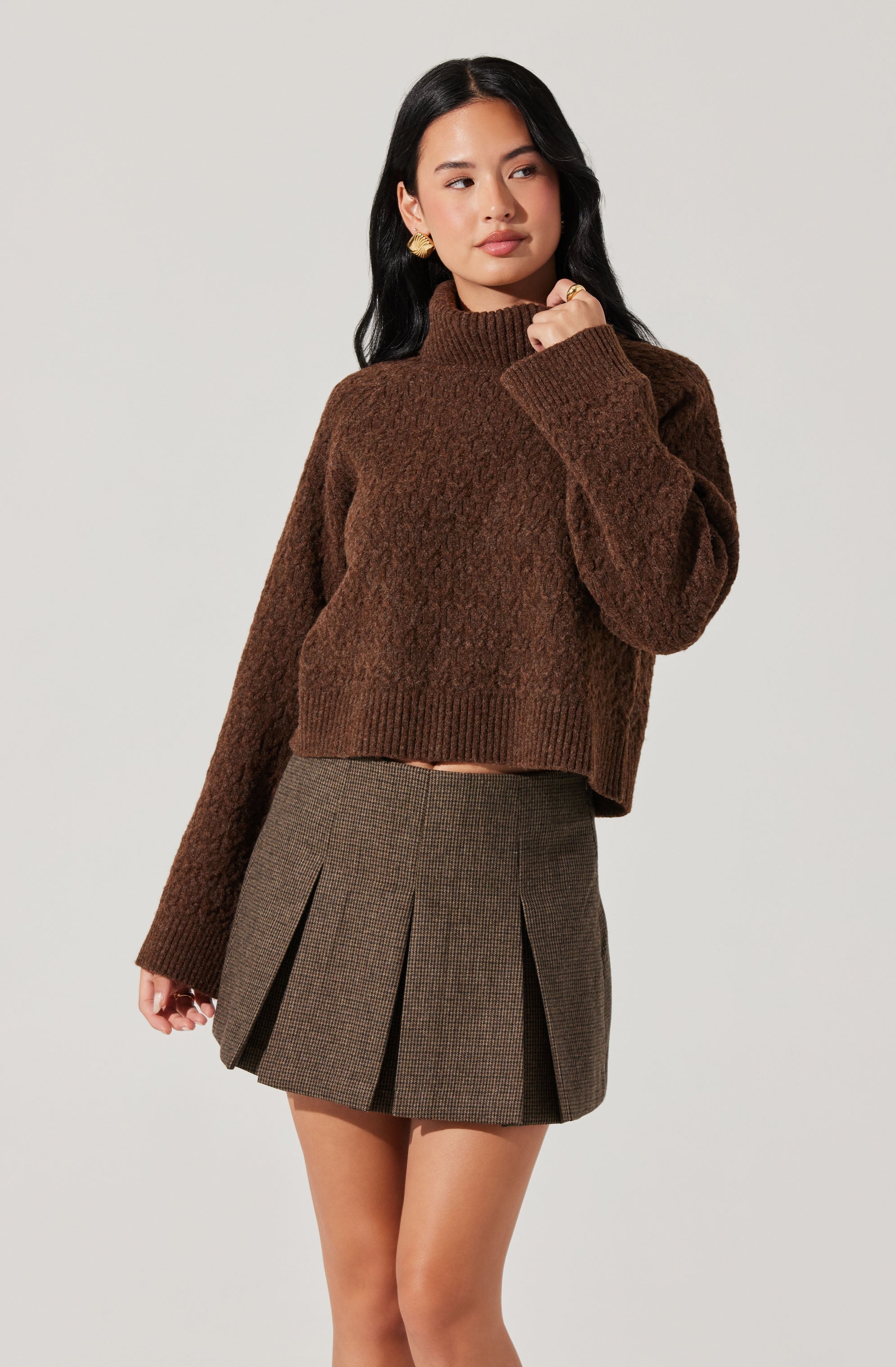 A woman is wearing a brown, chunky-knit turtleneck sweater with long sleeves and a slightly loose fit. She pairs it with a high-waisted, pleated mini skirt in a brown and black checkered pattern. She has long, dark hair, gold earrings, and a gold ring, and is posing with her left hand near her face against a plain, light-colored background.