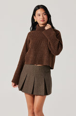 A woman is wearing a brown, chunky-knit turtleneck sweater with long sleeves and a slightly loose fit. She pairs it with a high-waisted, pleated mini skirt in a brown and black checkered pattern. She has long, dark hair, gold earrings, and a gold ring, and is posing with her left hand near her face against a plain, light-colored background.