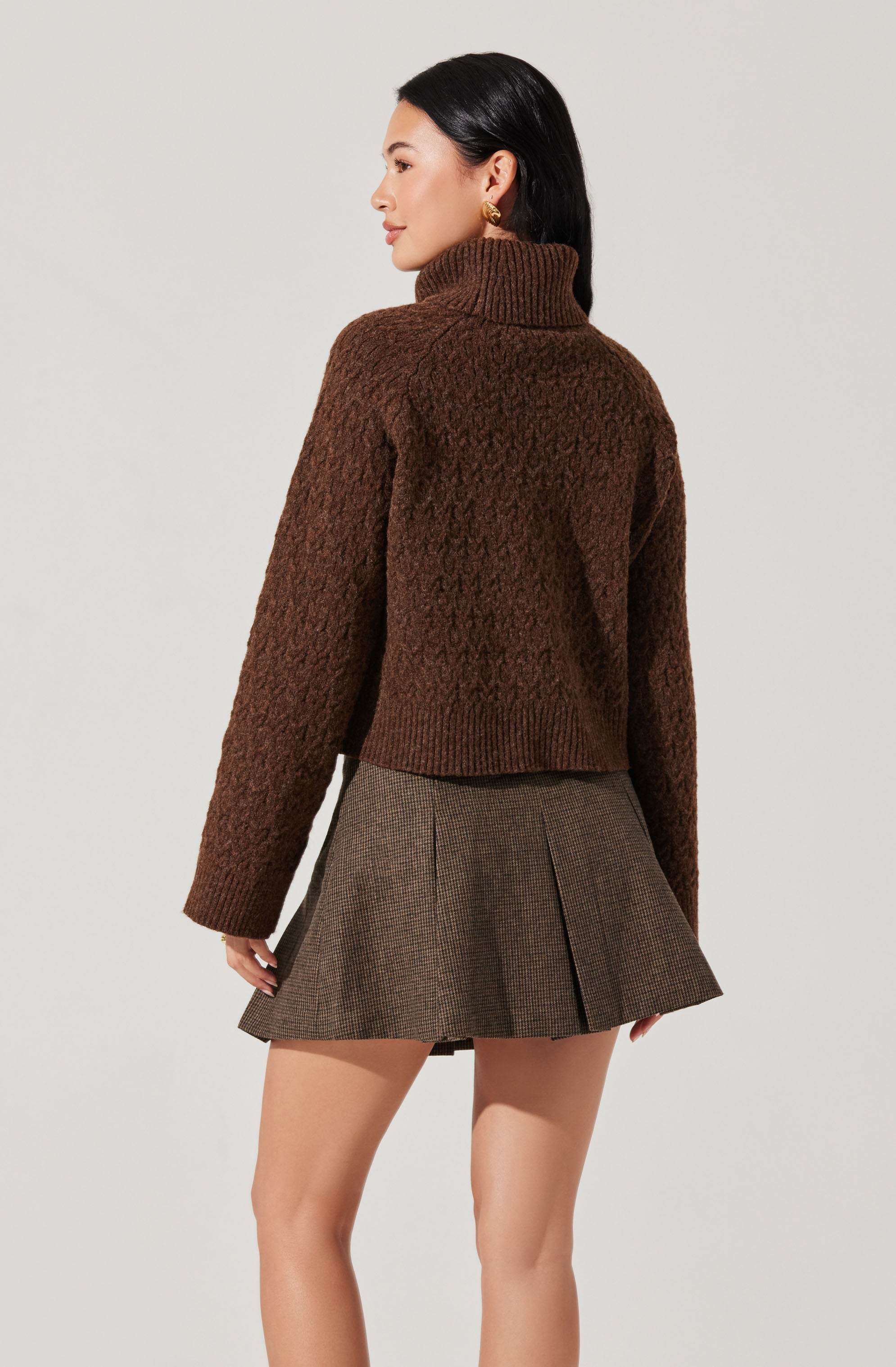 A woman is wearing a thick, textured brown turtleneck sweater with a ribbed collar. She is also wearing a high-waisted, pleated skirt with a brown and black checkered pattern.