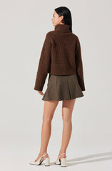 A woman is wearing a chunky brown turtleneck sweater with long sleeves and a ribbed hem. She pairs it with a high-waisted, pleated, plaid mini skirt in shades of brown and gray. She completes her outfit with white high-heeled shoes that have ankle straps.