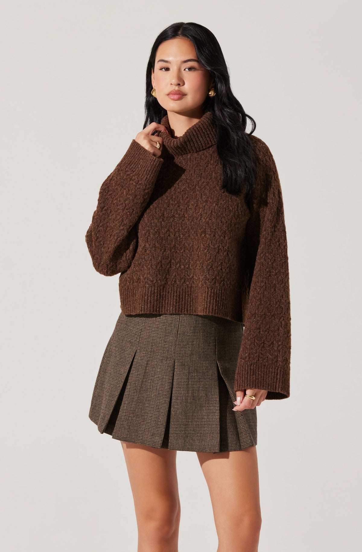 A woman is wearing a cozy, oversized brown turtleneck sweater with long sleeves, paired with a high-waisted, above-the-knee plaid skirt featuring pleats. The outfit has a classic, sophisticated style with earthy tones.