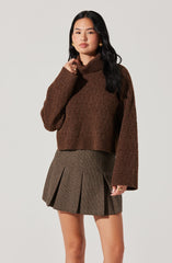 A woman is wearing a cozy, oversized brown turtleneck sweater with long sleeves, paired with a high-waisted, above-the-knee plaid skirt featuring pleats. The outfit has a classic, sophisticated style with earthy tones.
