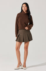 A woman is wearing a brown, chunky knit turtleneck sweater with long sleeves, paired with a high-waisted, pleated plaid skirt in shades of brown and gray. She is also wearing white high-heeled shoes with ankle straps.
