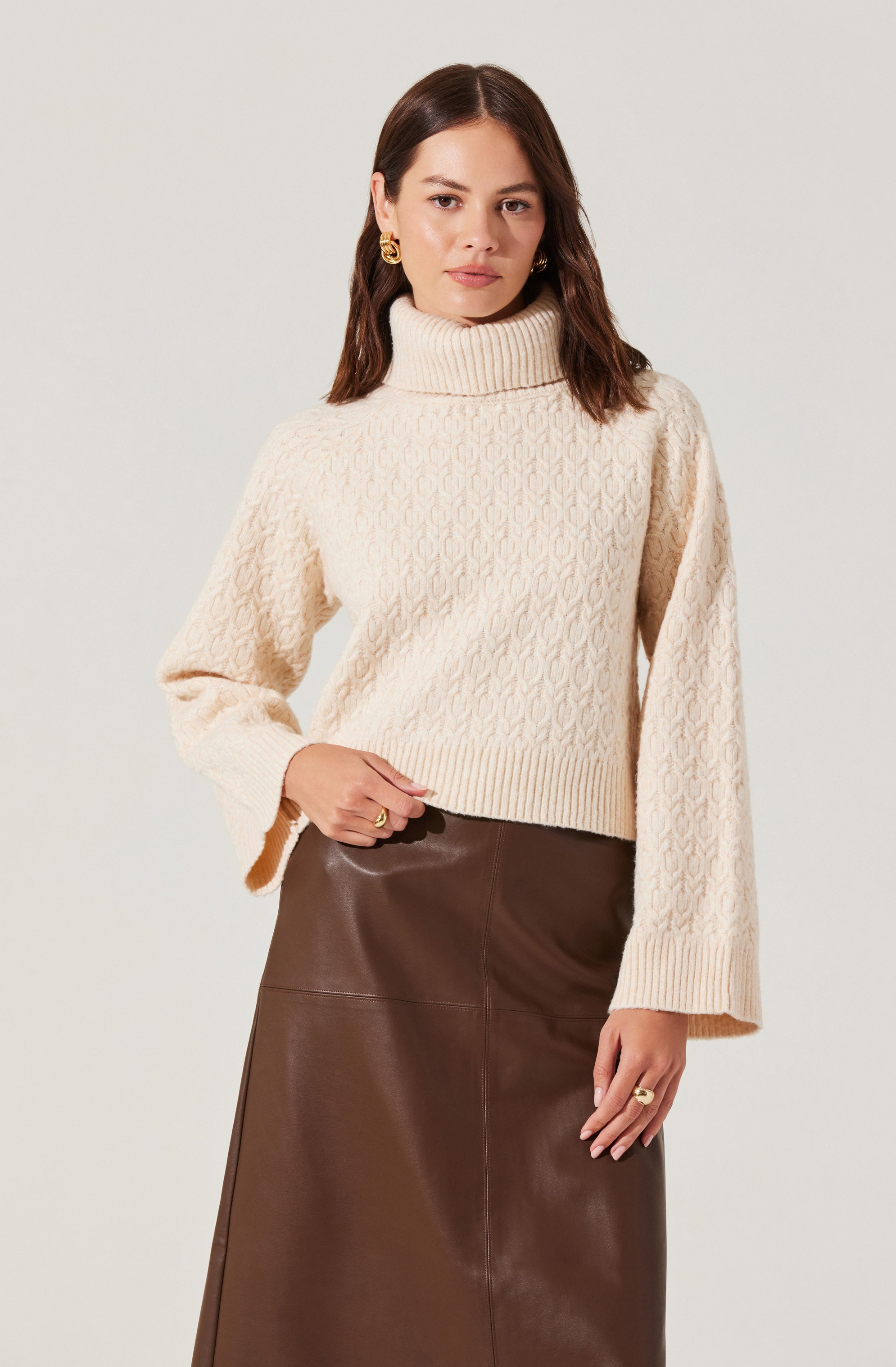 A woman is wearing a cream-colored, chunky cable-knit turtleneck sweater with long, slightly oversized sleeves. She pairs it with a high-waisted, dark brown leather skirt. She accessorizes with gold hoop earrings and gold rings on her fingers.