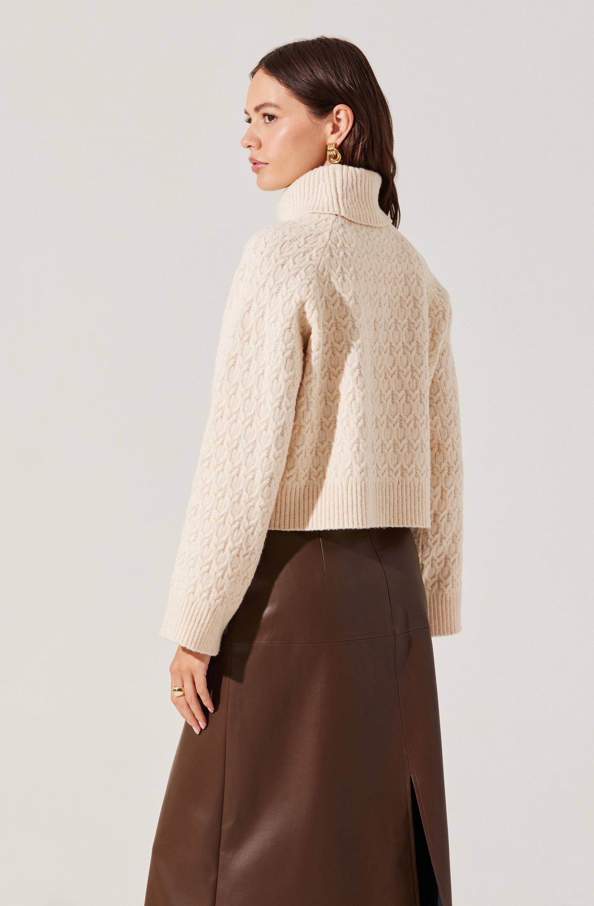 A woman is wearing a chunky cream-colored knit turtleneck sweater with a textured pattern and long sleeves. She pairs it with a high-waisted brown leather skirt featuring a front slit.