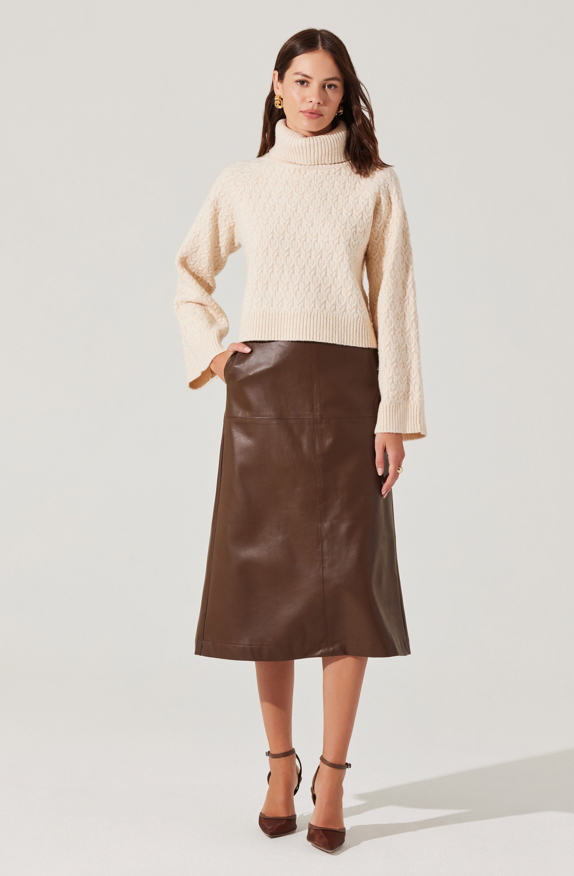 A woman is wearing a cream-colored, chunky knit turtleneck sweater with a cable pattern. She has paired it with a high-waisted, dark brown leather midi skirt that reaches below her knees. She is also wearing brown heels with ankle straps.