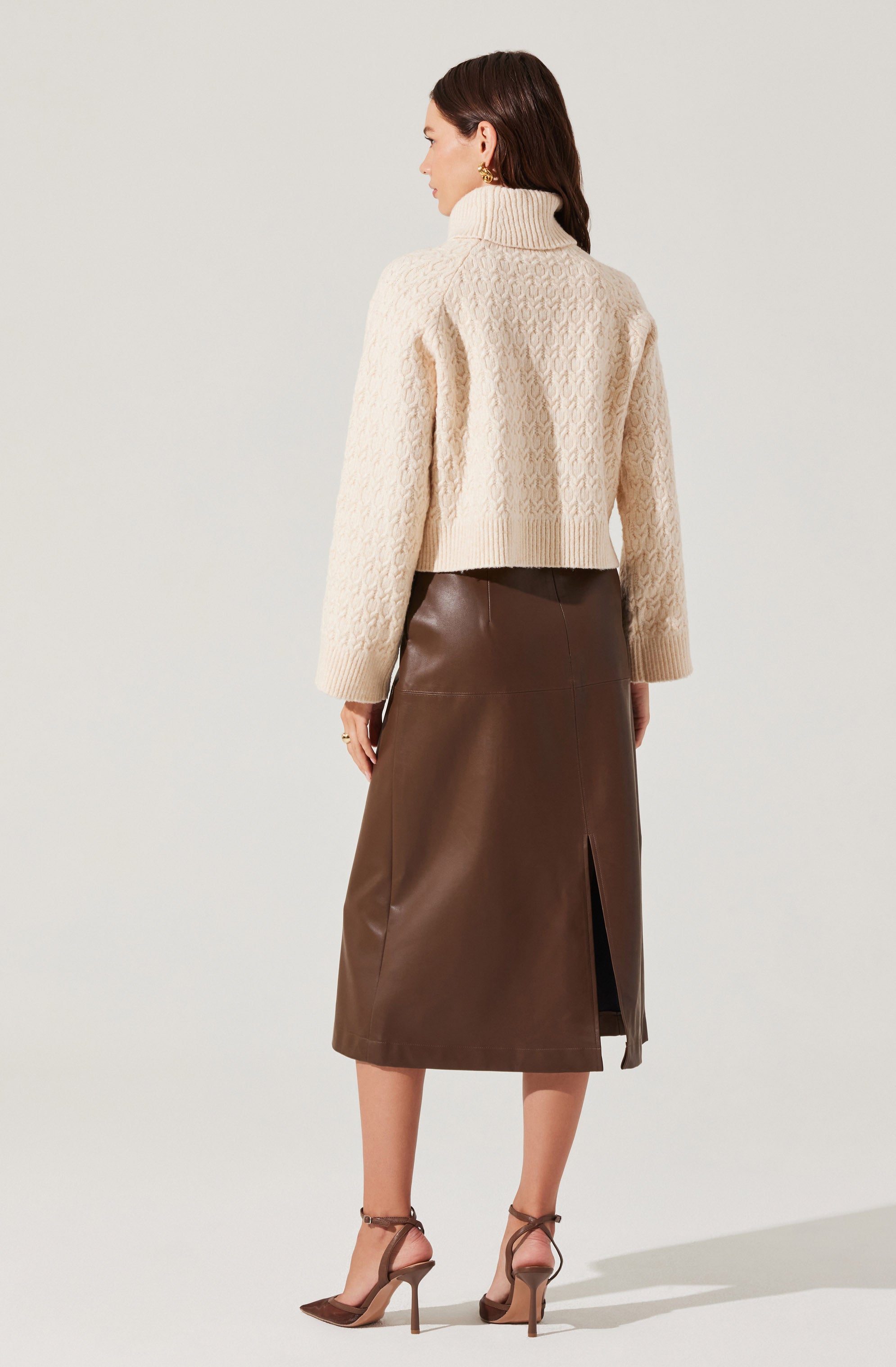 A woman is wearing a cream-colored, chunky knit turtleneck sweater with textured cable patterns. She pairs it with a high-waisted, brown leather midi skirt that features a front slit. She completes her outfit with strappy, pointed-toe, high-heeled shoes in a matching brown color.