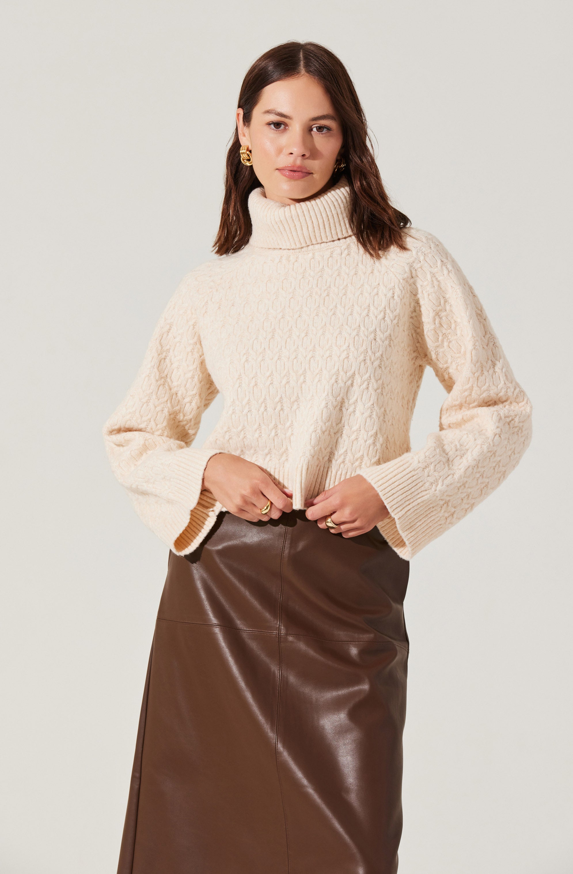 A woman is wearing a cream-colored, chunky knit turtleneck sweater with a textured pattern and oversized sleeves. She has paired it with a high-waisted brown leather skirt that reaches below her knees.