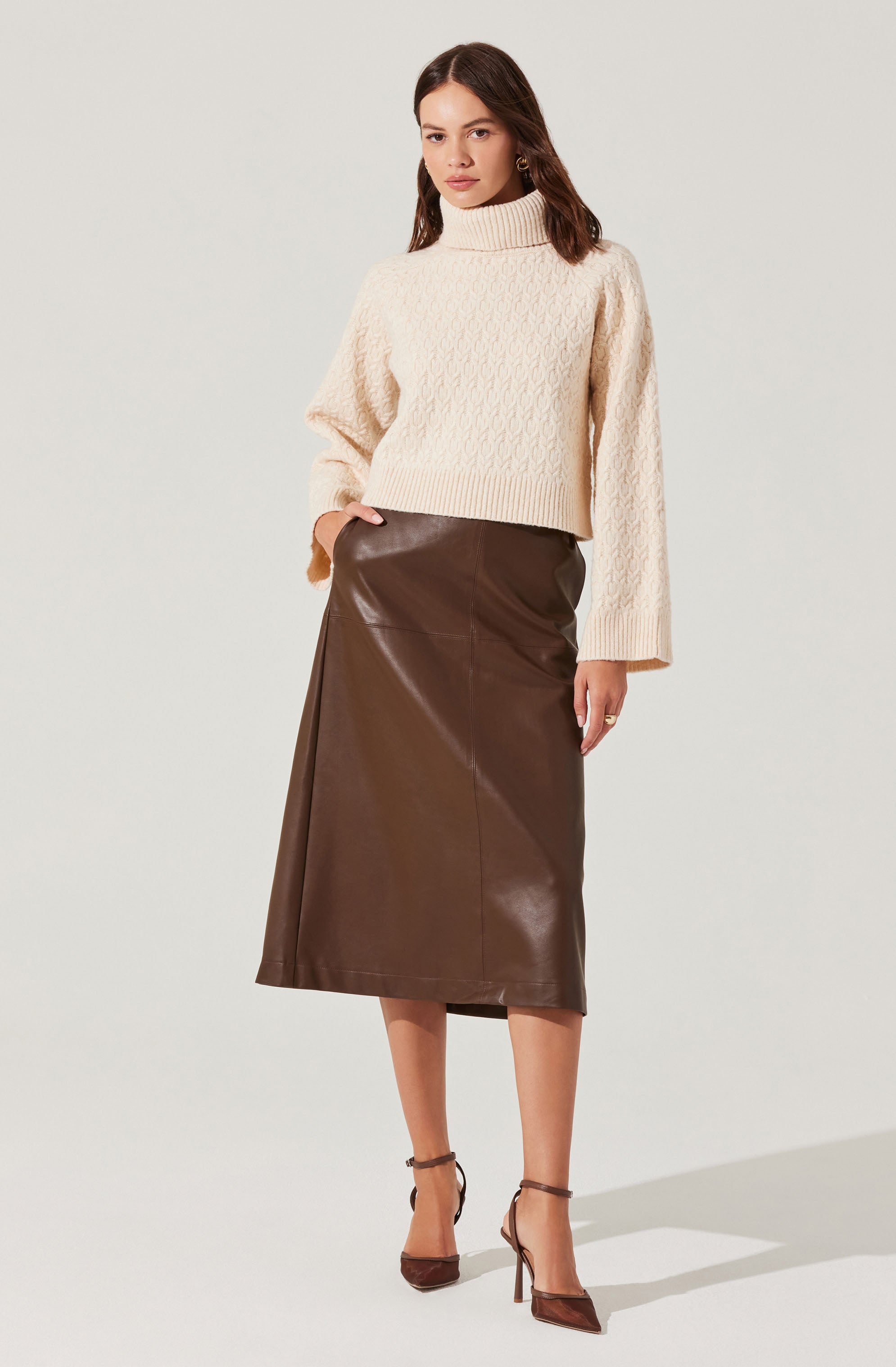 A woman is wearing a cream-colored chunky knit turtleneck sweater with long, loose sleeves. She pairs it with a high-waisted, brown leather midi skirt that falls below her knees. She completes the outfit with brown high-heeled shoes with ankle straps.