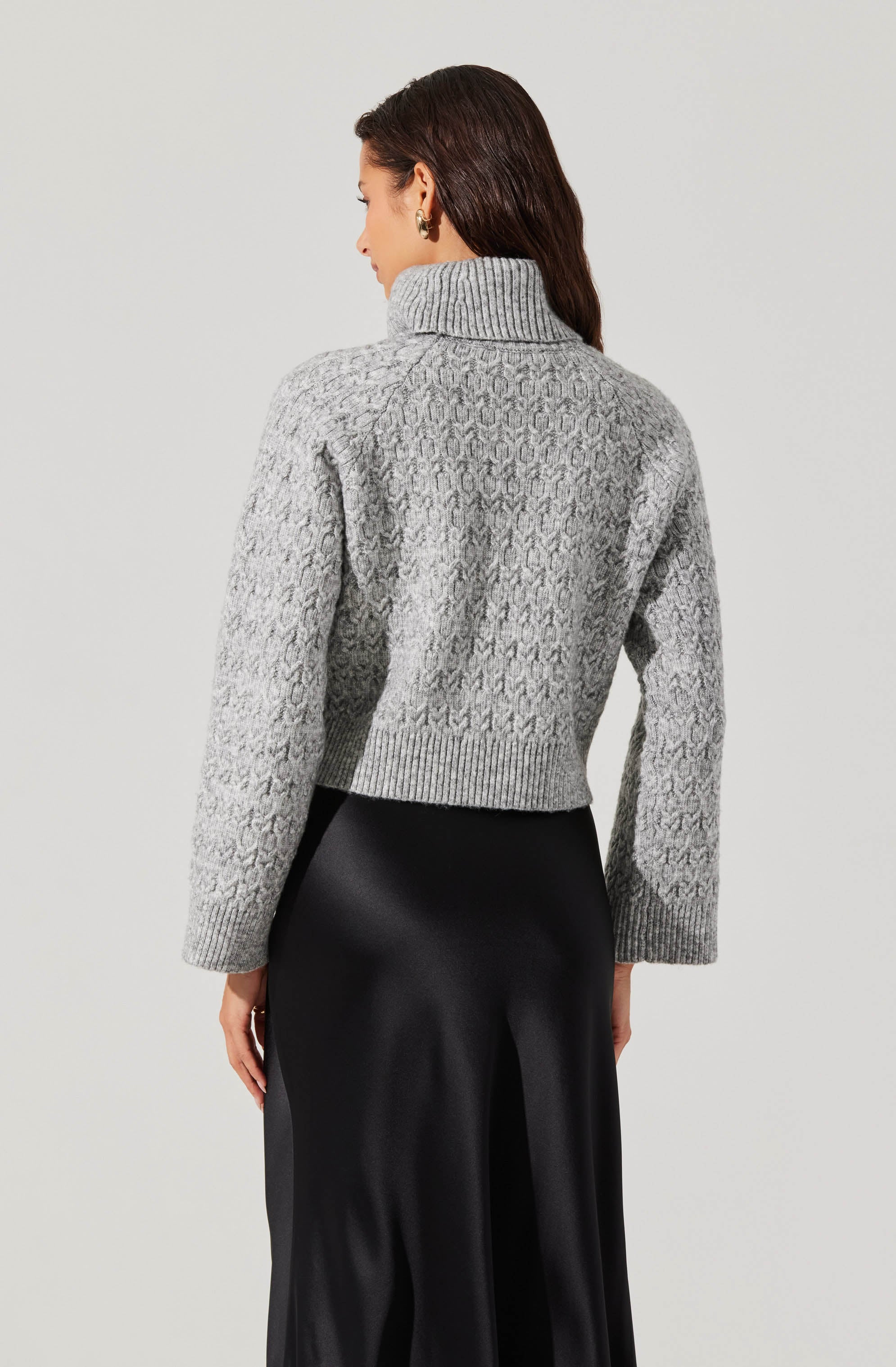 A woman is wearing a gray, chunky knit turtleneck sweater with a textured pattern and wide ribbed cuffs. She pairs it with a black satin skirt that has a smooth, shiny finish and a flowing silhouette.