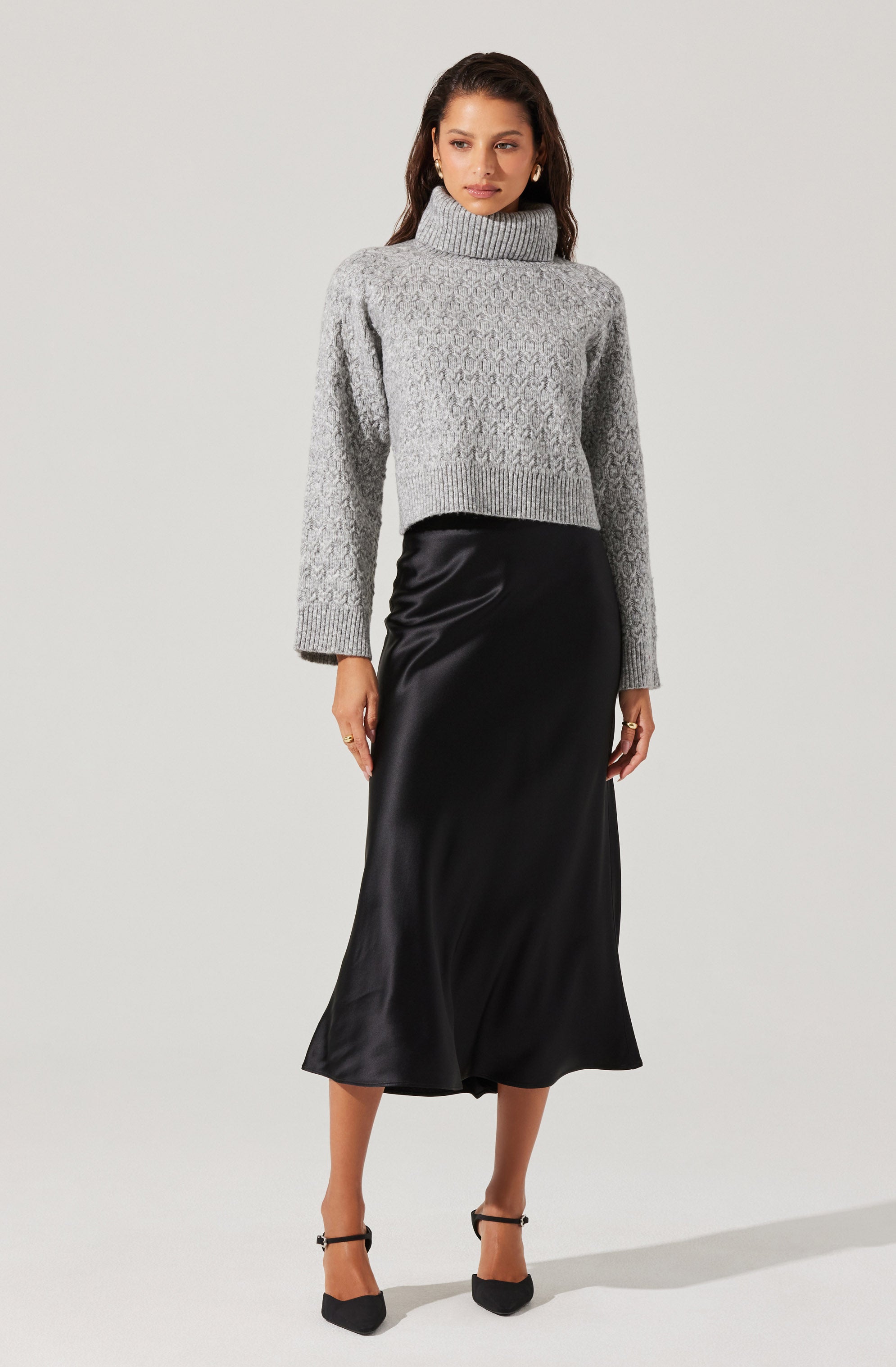 A woman is wearing a gray knit turtleneck sweater with a textured pattern and long sleeves. She pairs it with a black satin midi skirt that has a slight sheen and falls below the knees. She completes her outfit with black pointed-toe heels.