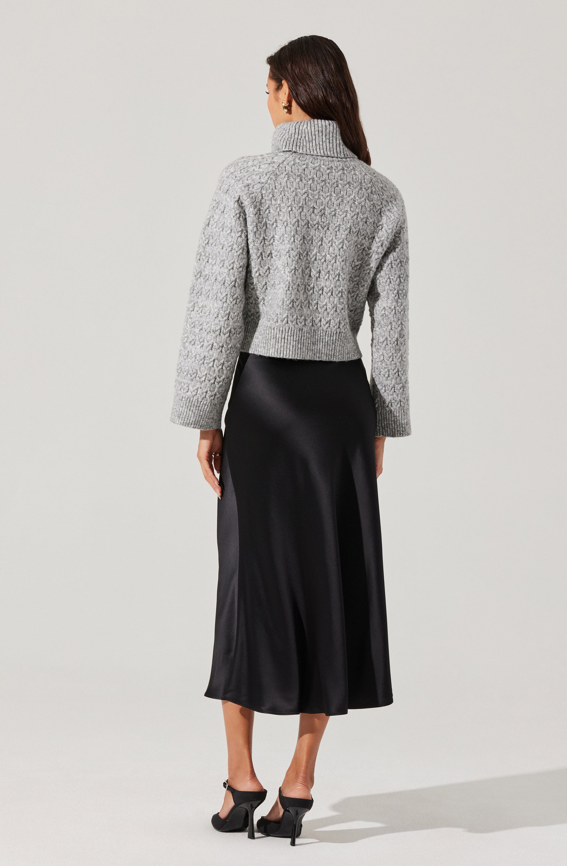 A woman is wearing a textured, light gray, chunky knit sweater with a wide collar and long sleeves. She pairs it with a black satin midi skirt that has a smooth, shiny surface and a flowy silhouette. She is also wearing black high-heeled open-toe sandals.