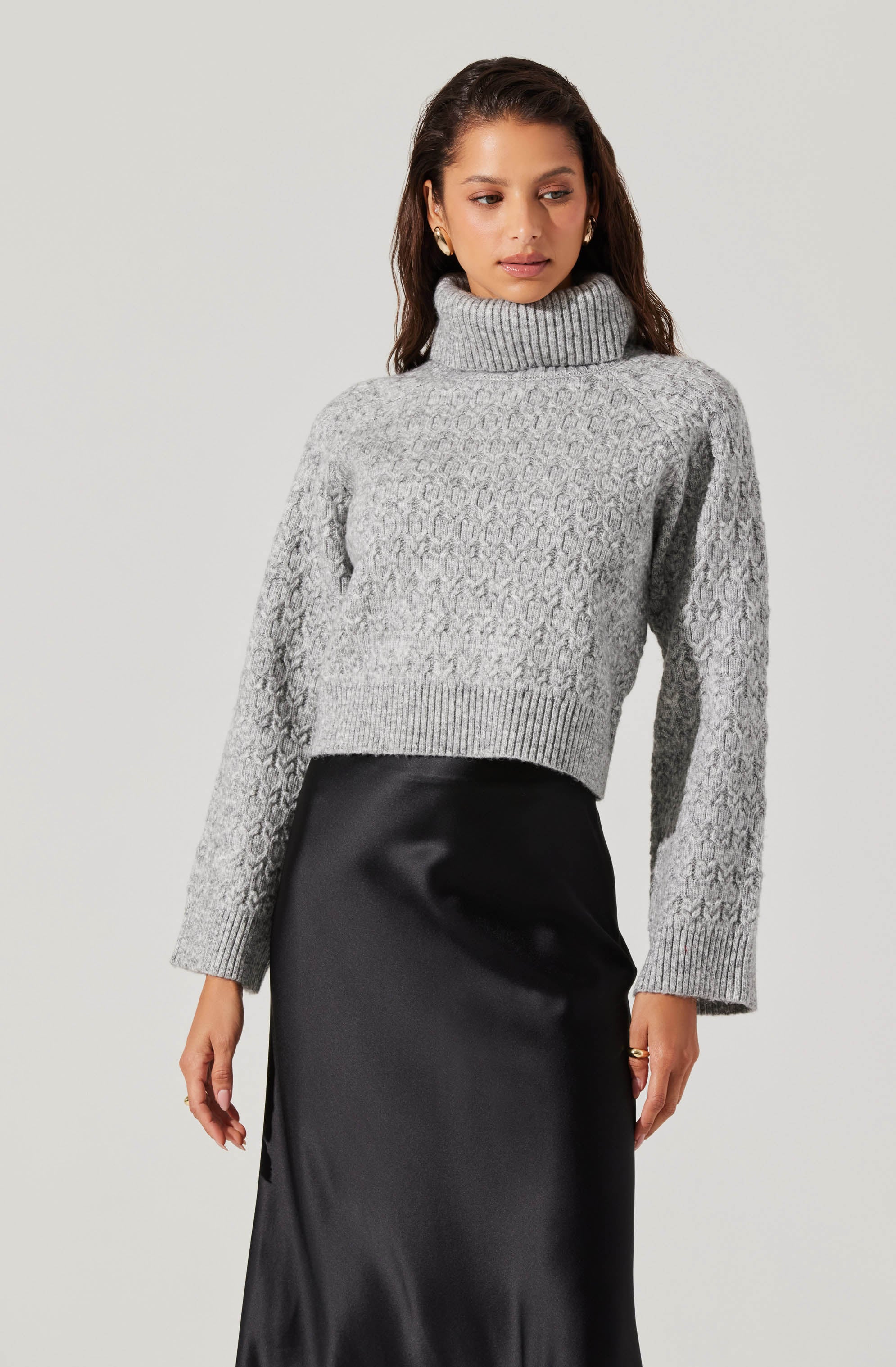 A woman is wearing a light gray, chunky-knit turtleneck sweater with a textured cable pattern and ribbed cuffs and hem. She pairs it with a high-waisted, black satin skirt.