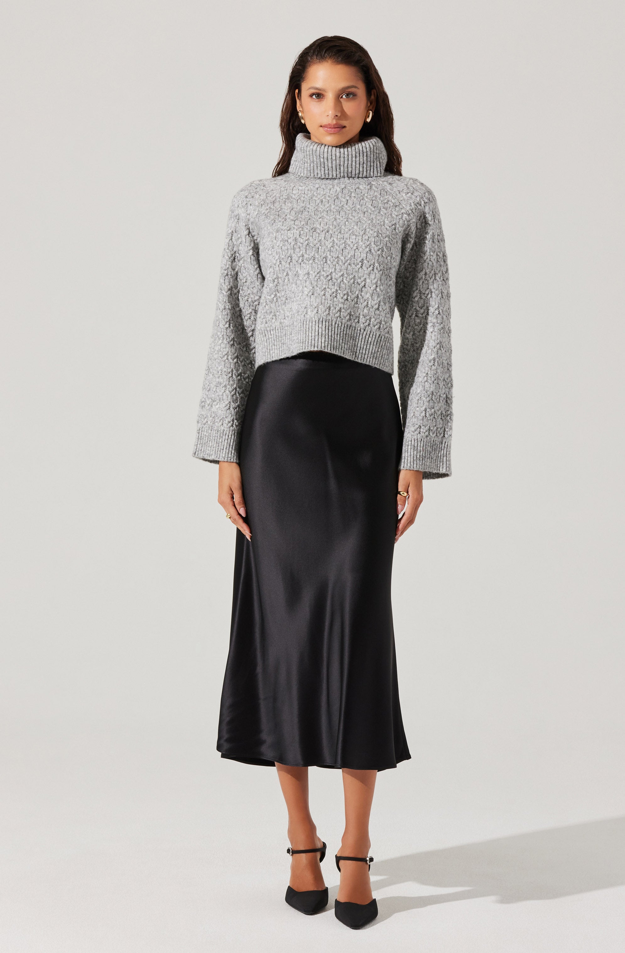 A woman is wearing a light gray, chunky knit turtleneck sweater with long sleeves. She pairs it with a high-waisted, black satin midi skirt that reaches mid-calf. She completes the outfit with black pointed-toe heels with ankle straps.