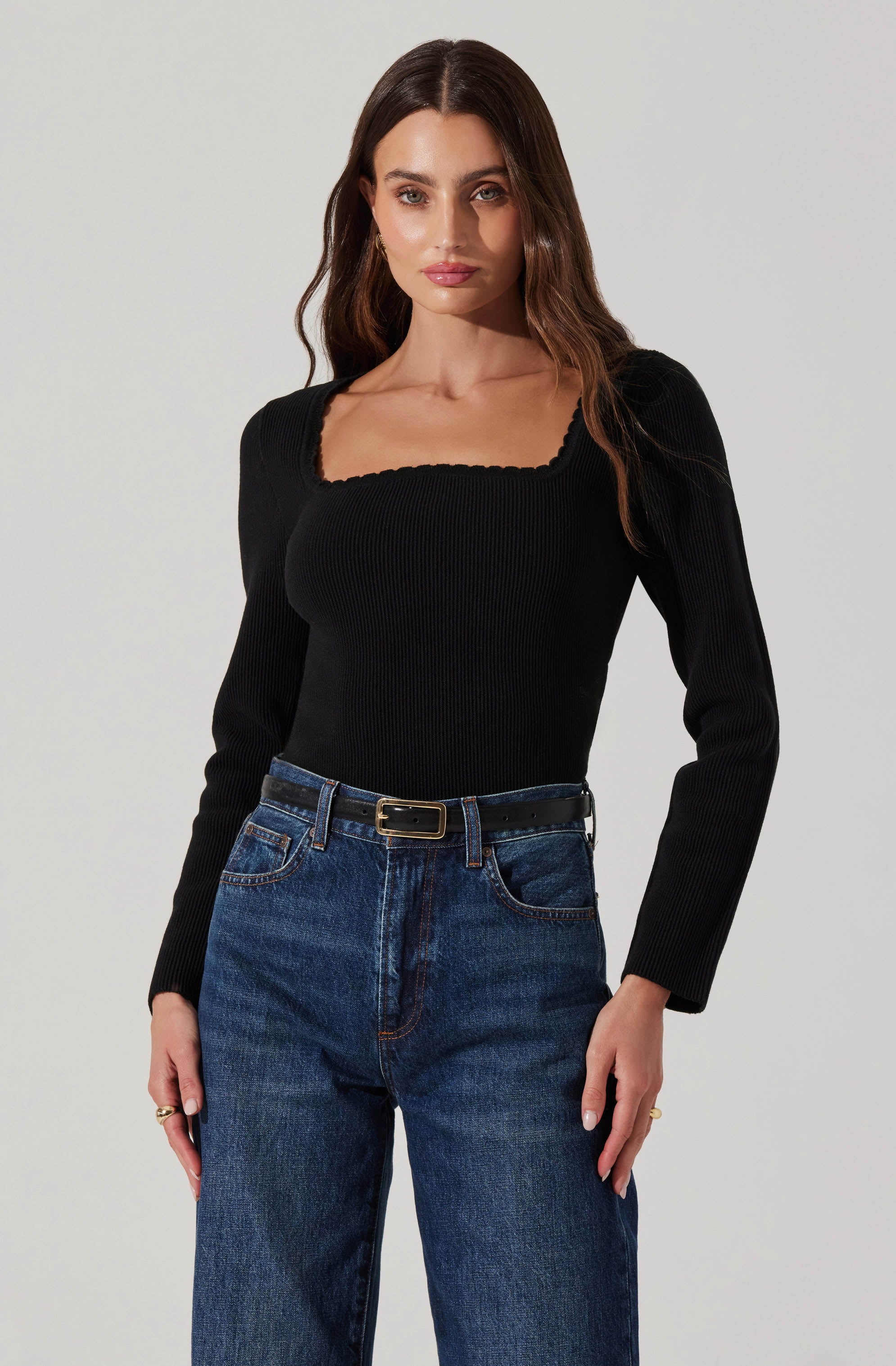 A woman is wearing a fitted black long-sleeve top with a scalloped square neckline and ribbed texture, paired with high-waisted blue jeans with a relaxed fit, secured with a black belt featuring a gold buckle.