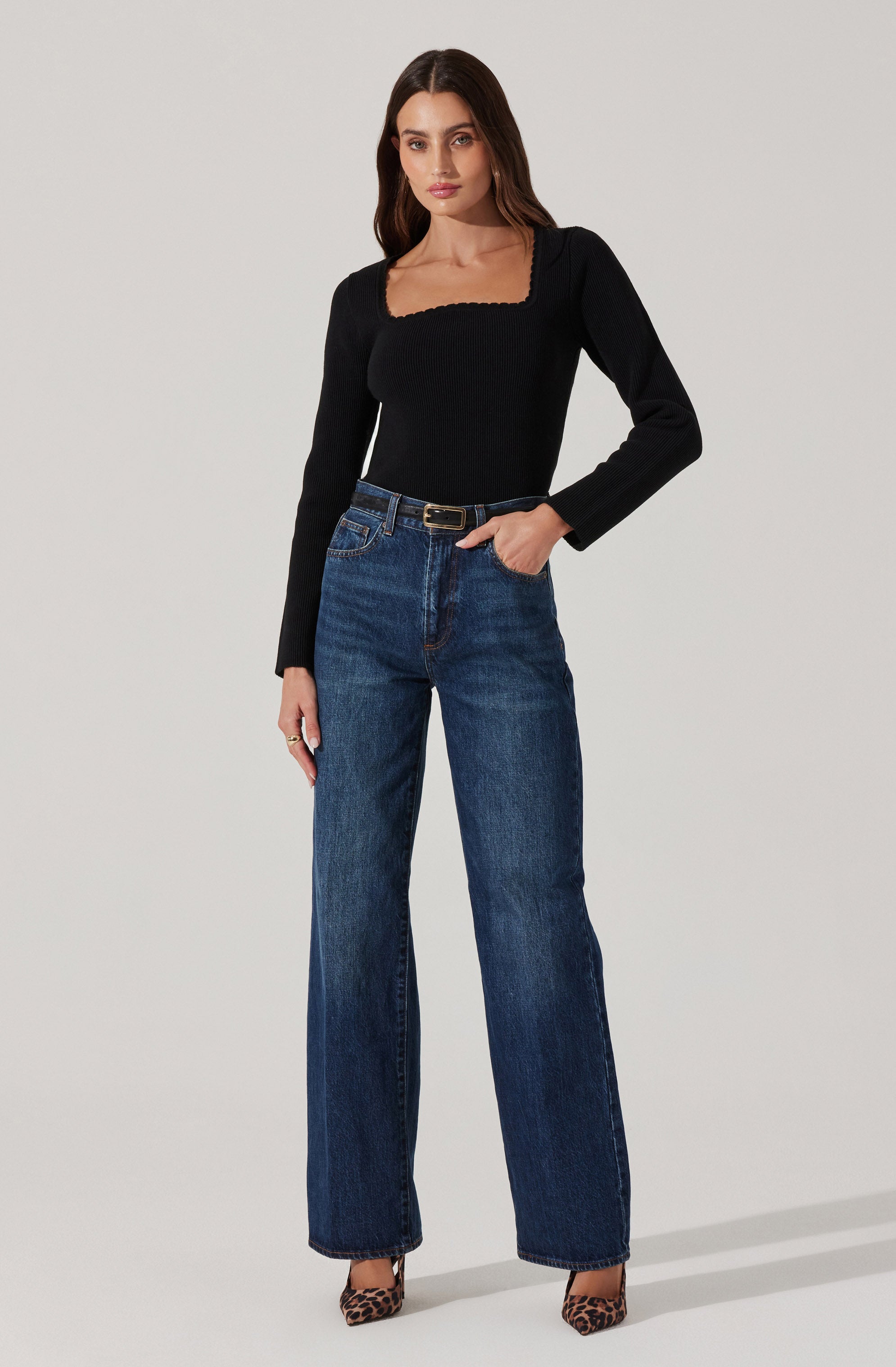 A woman is wearing a fitted black long-sleeve top with a square neckline and scalloped edges, paired with high-waisted, straight-leg blue jeans. She completes the look with a black belt that has a gold buckle and leopard-print high heels.