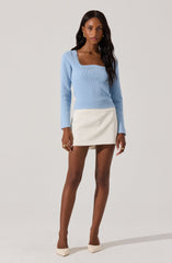 A woman is wearing a light blue long-sleeve sweater with a square neckline and scalloped edges. She pairs it with a high-waisted, white A-line mini skirt. She completes her outfit with light-colored closed-toe heels and multiple gold rings and earrings.