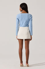 A woman is wearing a light blue long-sleeve sweater with a round neckline and a white mini skirt with a slight A-line shape and clean lines. She is also wearing nude-colored high-heeled shoes.