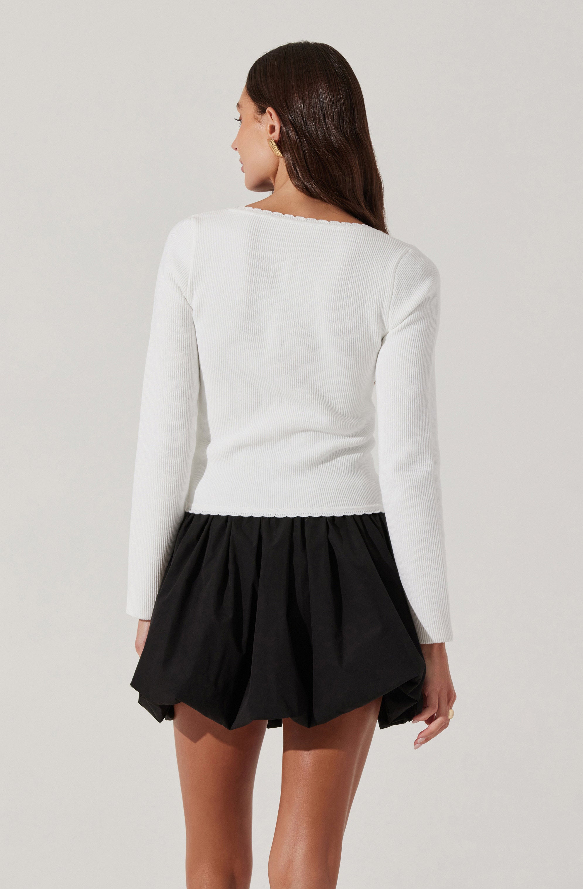 A woman is wearing a long-sleeved, white ribbed sweater with a slightly scalloped neckline. She pairs it with a short, black, puffed skirt that has a gathered design, creating a voluminous appearance.