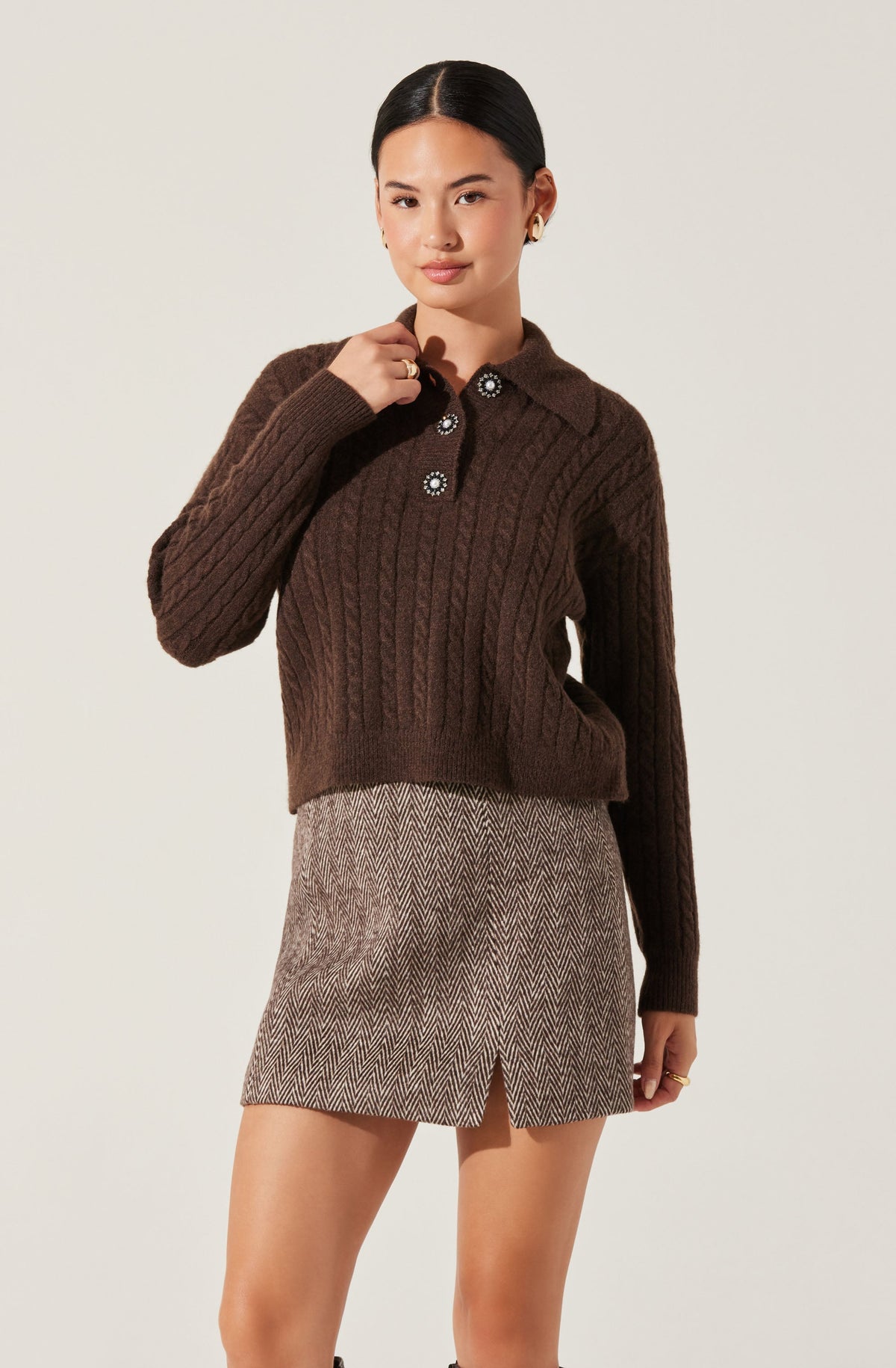 A woman is wearing a dark brown, chunky knit sweater with a cable pattern. The sweater features a collared neckline with four decorative buttons, each adorned with a flower-like design, and long sleeves. She also wears a beige and brown herringbone patterned skirt with a small slit in the front.
