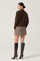 A woman is wearing a dark brown, cable-knit sweater with a high collar and long sleeves. She pairs it with a fitted, herringbone-patterned mini skirt in neutral tones. She completes her outfit with knee-high, black leather stiletto boots.