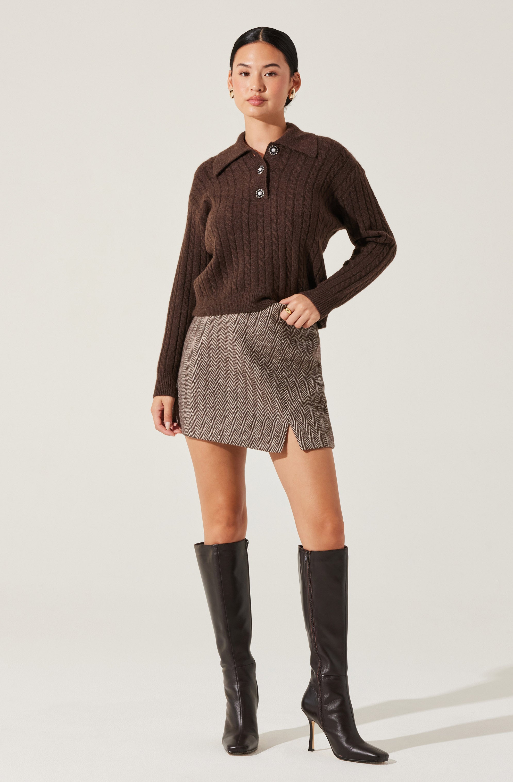 A woman is wearing a dark brown, long-sleeve, ribbed sweater with a collar and three buttons at the neckline. She pairs it with a high-waisted, plaid mini skirt that has a small slit on the front. She completes her outfit with black, knee-high, pointed-toe boots with high heels.