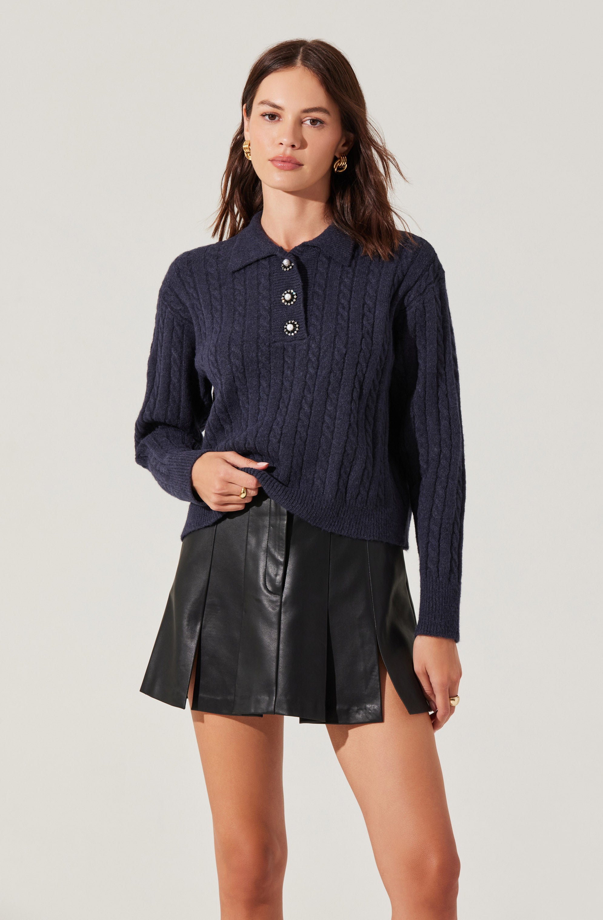 A woman is wearing a navy blue, cable-knit sweater with a collared neckline and three decorative pearl and rhinestone buttons down the front. She pairs it with a black, faux leather mini skirt featuring vertical panels and side slits.