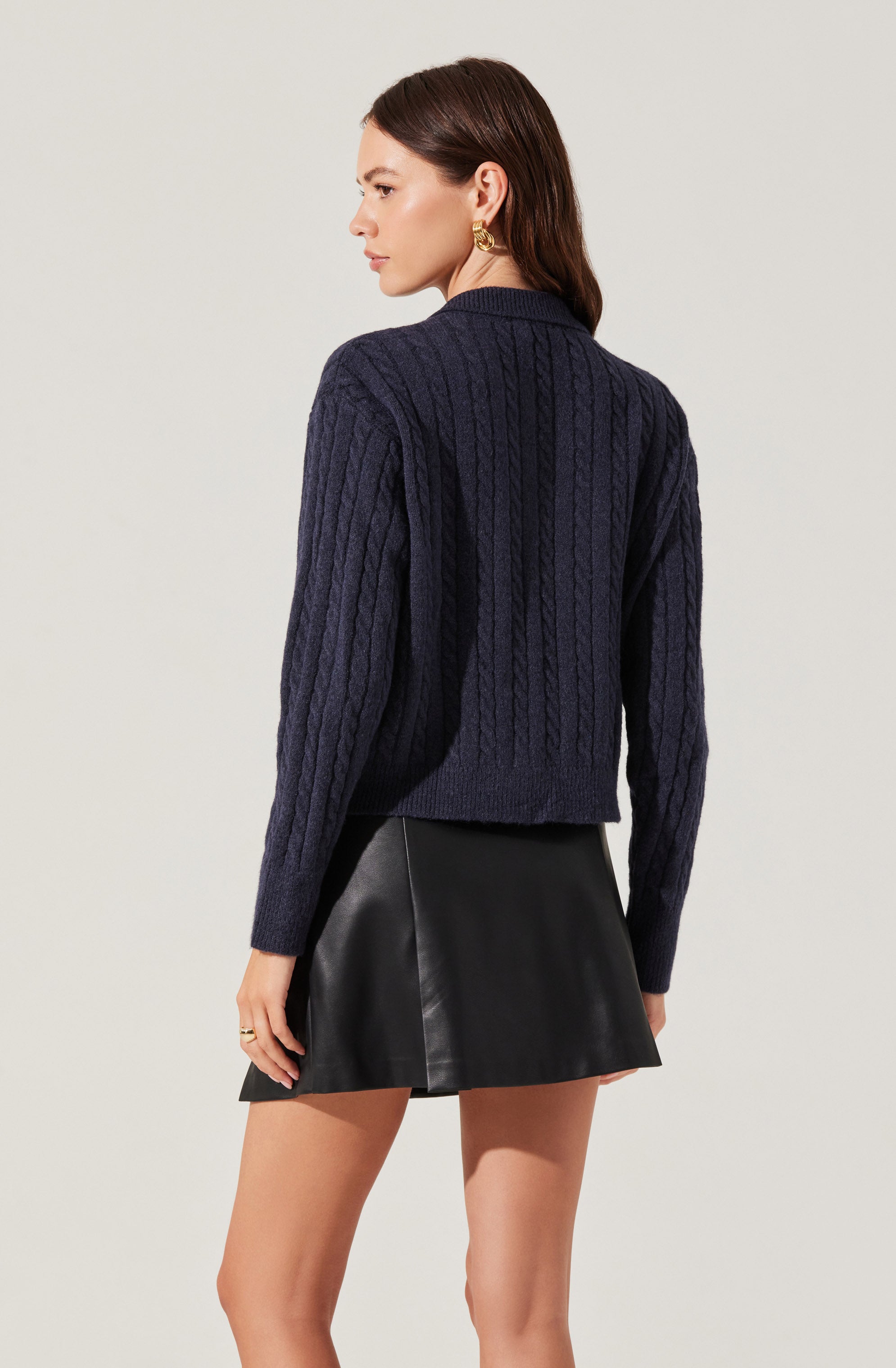 A woman is wearing a dark navy blue cable-knit sweater with a textured pattern and a loose fit. She is also wearing a black leather mini skirt with an A-line silhouette. The outfit is accessorized with gold hoop earrings.