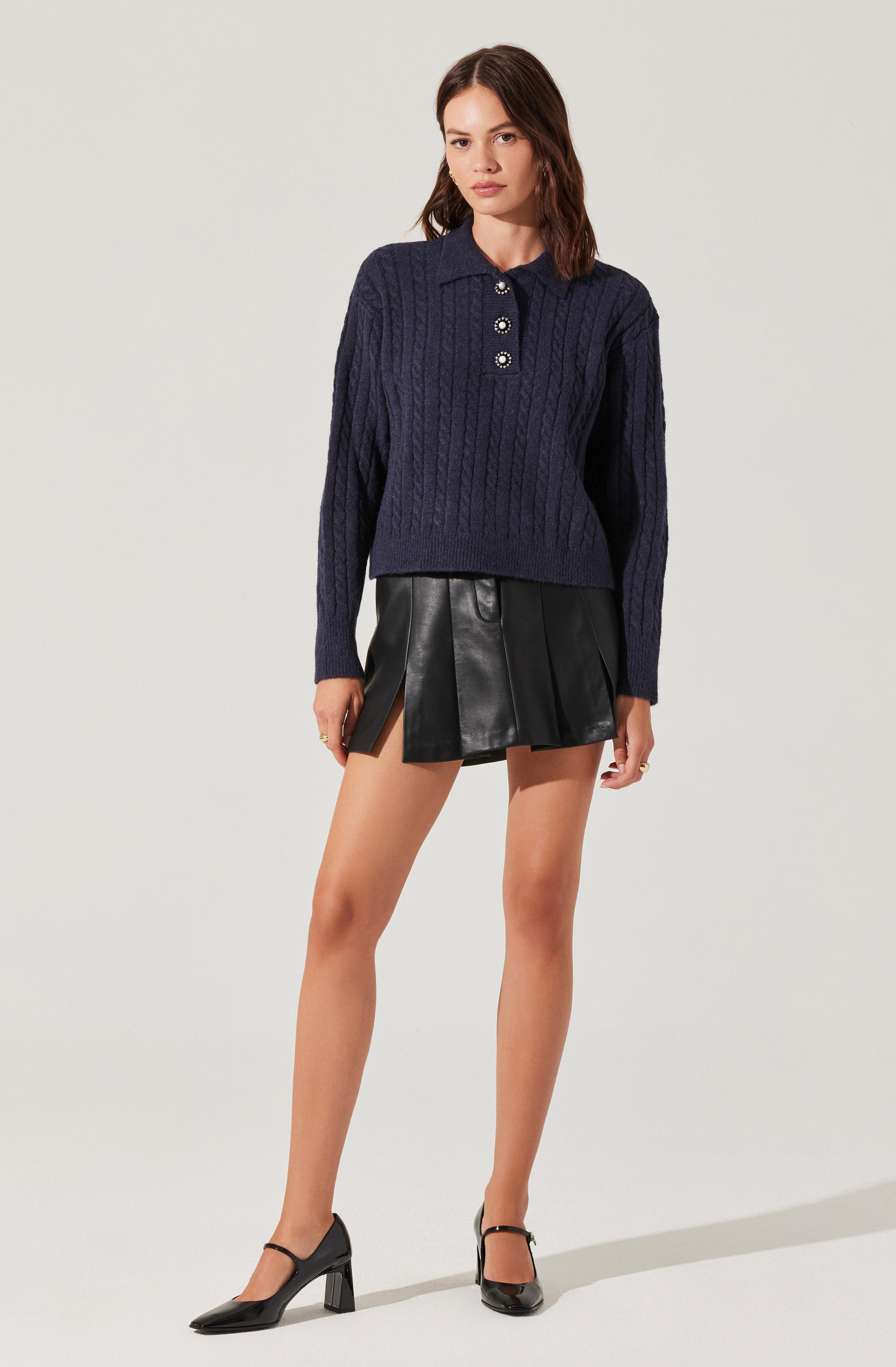 A woman is wearing a navy blue, long-sleeved, cable-knit sweater with a collar and button placket. She pairs it with a black leather mini skirt that features a pleated design and an asymmetrical hem. She completes the outfit with black, closed-toe, ankle-strap high heels.