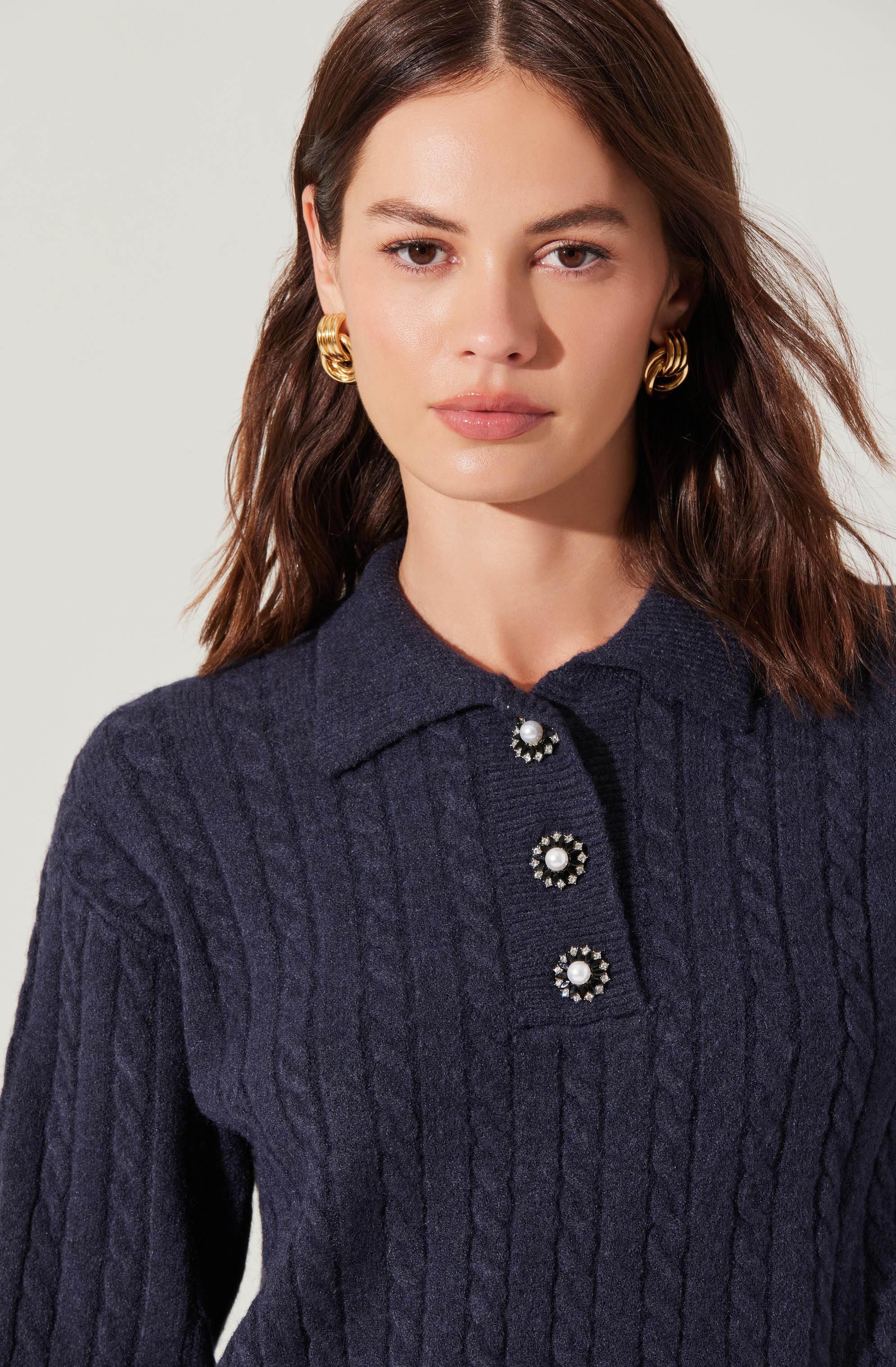 A woman is wearing a dark navy blue cable-knit sweater with a collared neckline and a button placket featuring three decorative buttons adorned with pearls and rhinestones. She has medium-length brown hair and is accessorized with large, gold hoop earrings.
