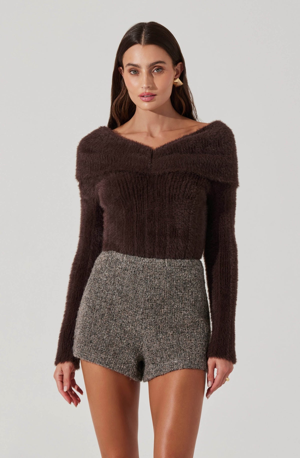 A woman is wearing a dark brown, long-sleeved, fuzzy sweater with a wide neckline that drapes off the shoulders. She is also wearing high-waisted, gray, textured shorts.