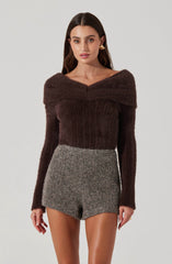 A woman is wearing a dark brown, long-sleeved, fuzzy sweater with a wide neckline that drapes off the shoulders. She is also wearing high-waisted, gray, textured shorts.