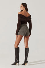A woman is wearing an off-the-shoulder, long-sleeve, fuzzy brown sweater paired with high-waisted, short, gray tweed shorts. She has on tall black leather high-heeled boots. She is also accessorized with a large gold earring.