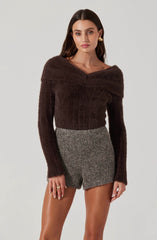 A woman is wearing a dark brown fuzzy knit sweater with a wide, off-the-shoulder neckline and long sleeves. She pairs it with high-waisted, gray tweed shorts.