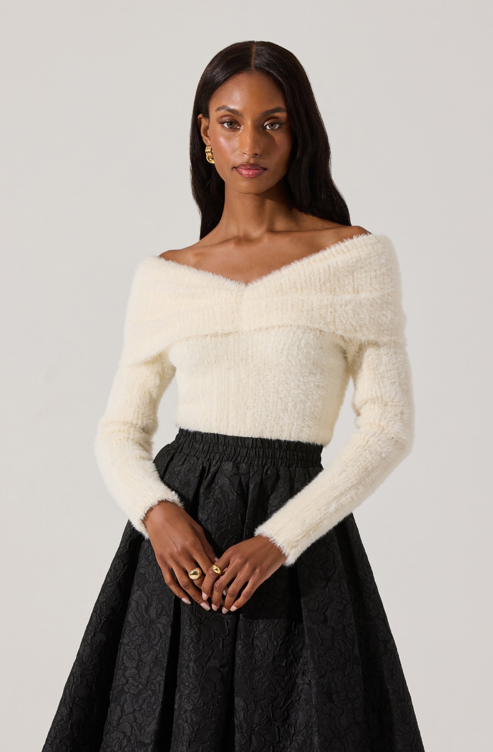 A woman is wearing a white, off-the-shoulder, fuzzy sweater with a wide neckline that drapes over her shoulders. She pairs it with a high-waisted, black skirt with a subtle floral pattern and gathered waistband. She accessorizes with gold earrings and rings.