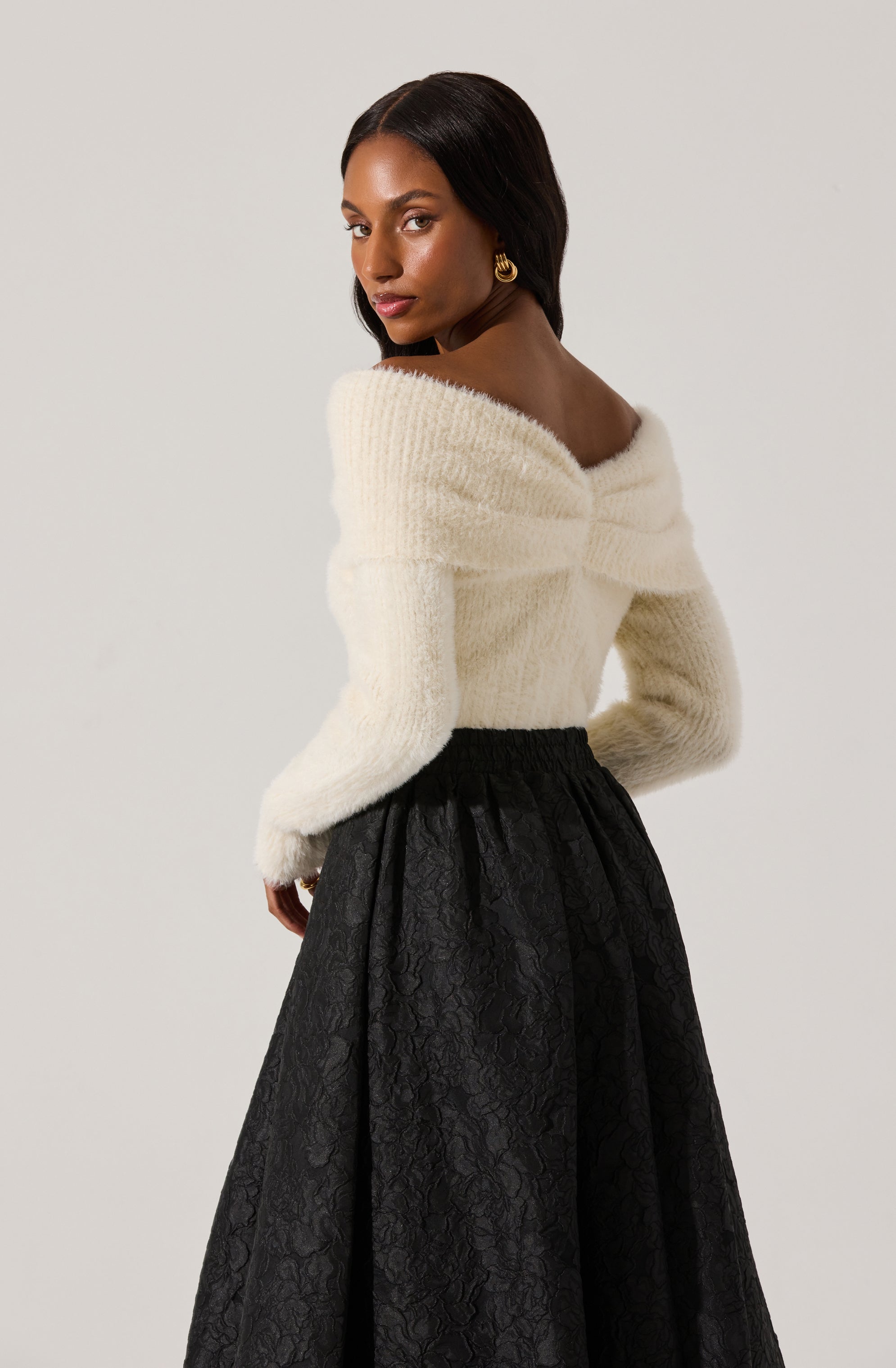 A woman is wearing a soft, off-the-shoulder, cream-colored fuzzy sweater with long sleeves. She is paired it with a high-waisted, black, full-length skirt featuring an intricate lace pattern.