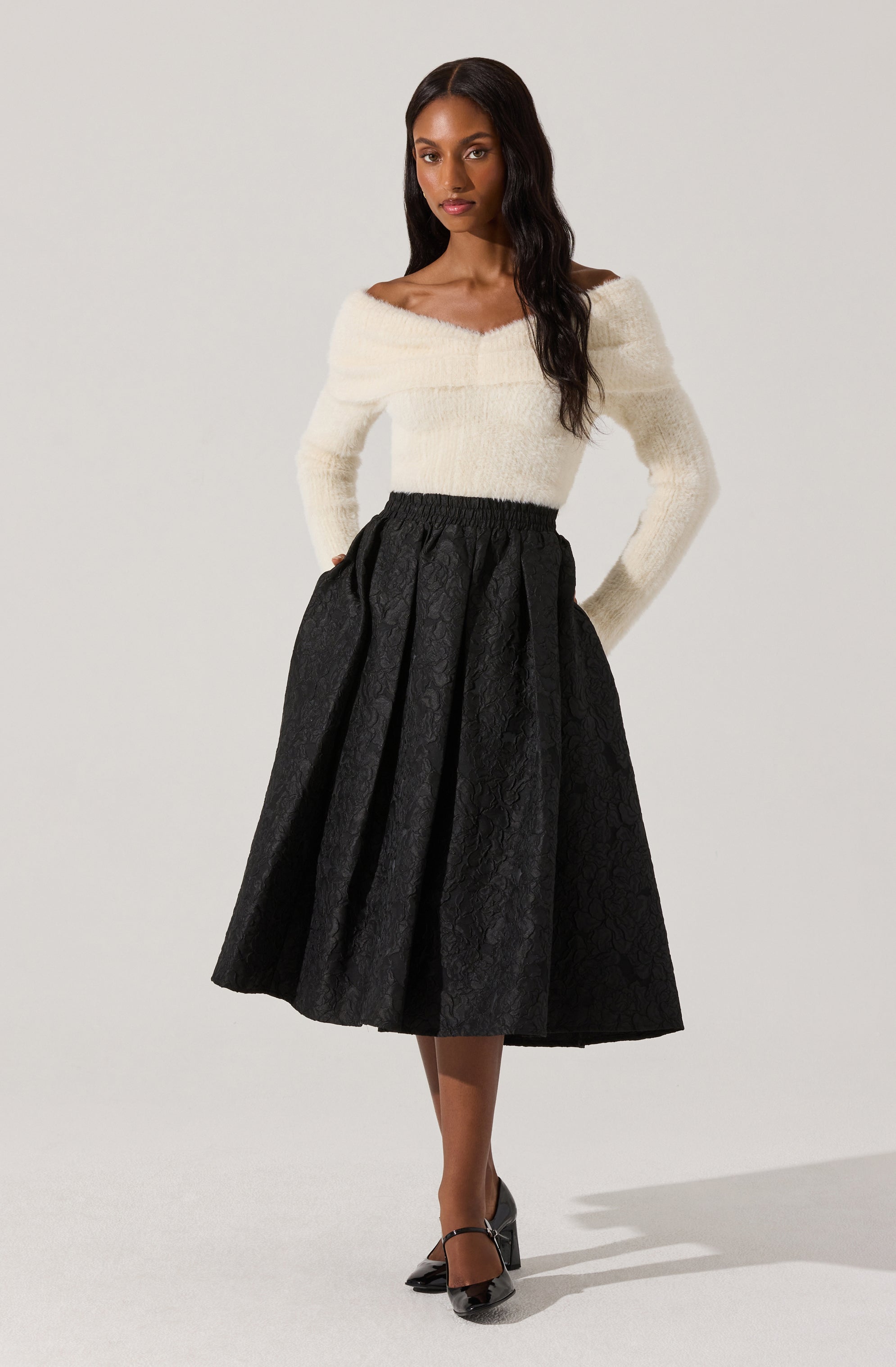The woman is wearing a soft, off-the-shoulder white sweater with a fuzzy texture, paired with a high-waisted, black, midi-length skirt that has a subtle floral pattern. She is also wearing black, pointed-toe heels with ankle straps.