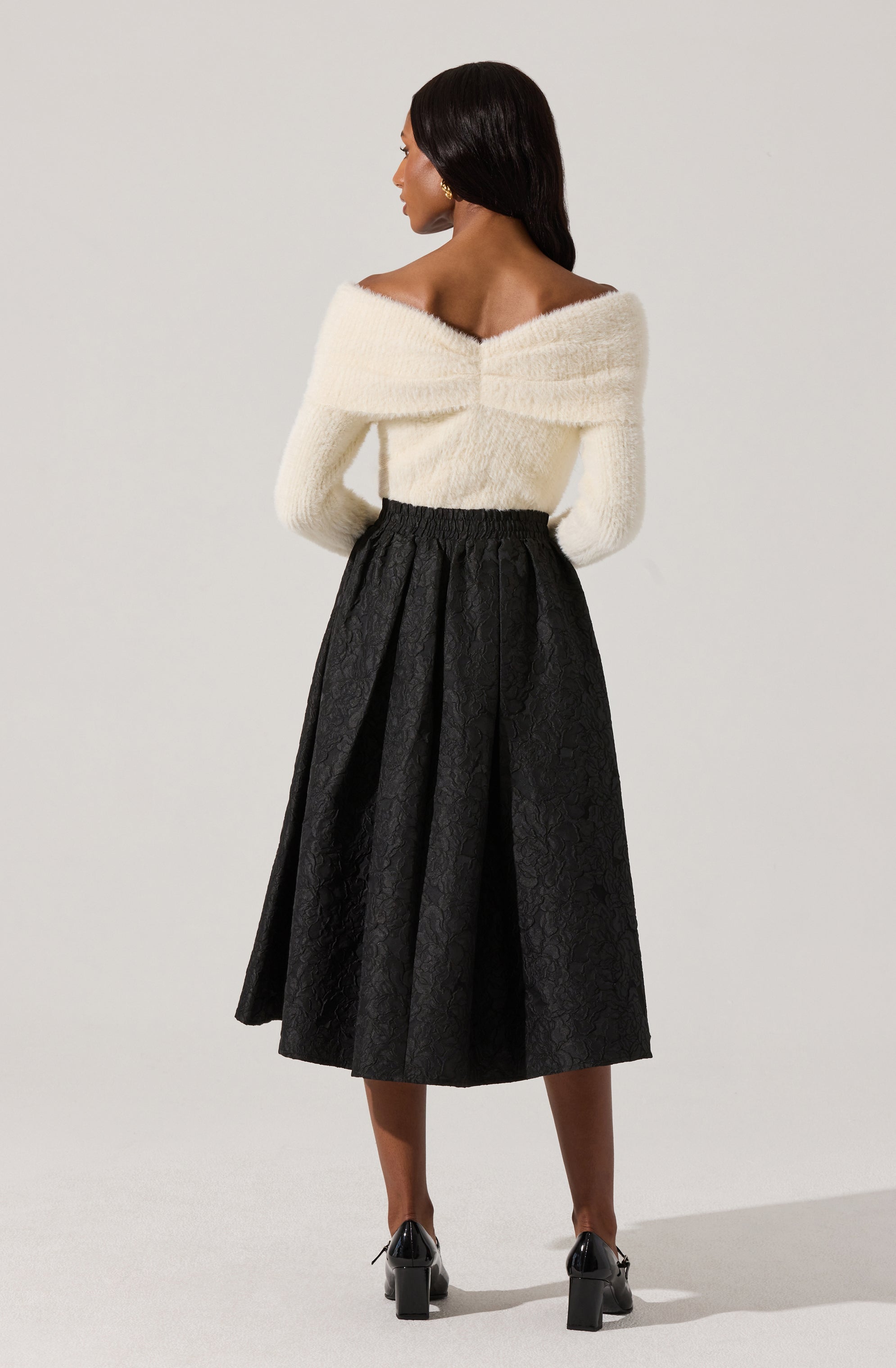 A woman is wearing an off-the-shoulder, long-sleeved, white fuzzy sweater paired with a high-waisted, black midi skirt that has a textured, patterned fabric. She is also wearing black heeled shoes.