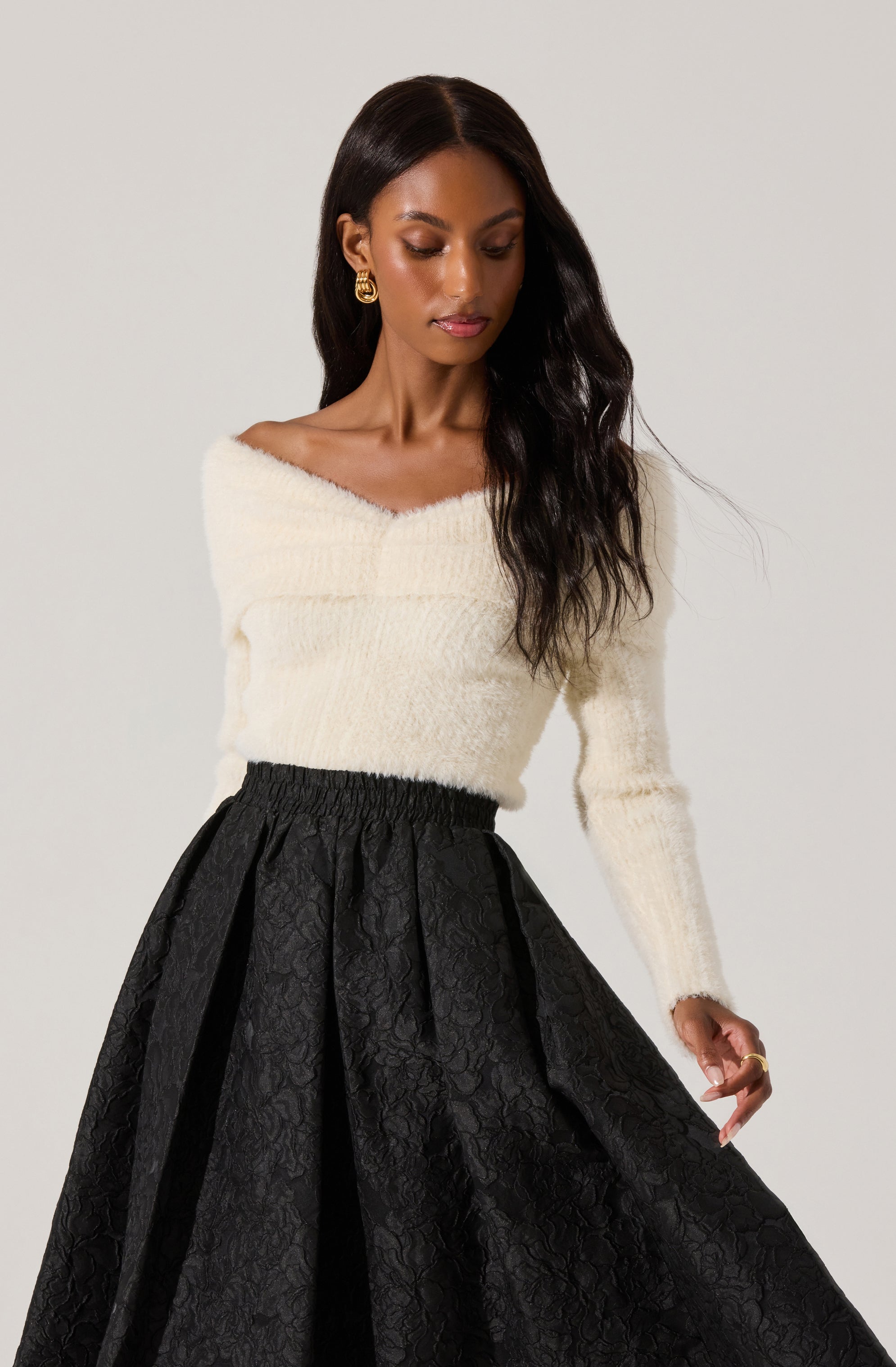 A woman is wearing a soft, off-the-shoulder, cream-colored fuzzy sweater with long sleeves. She pairs it with a high-waisted, full black skirt that has a textured, floral pattern and pleats, creating an elegant and sophisticated look.
