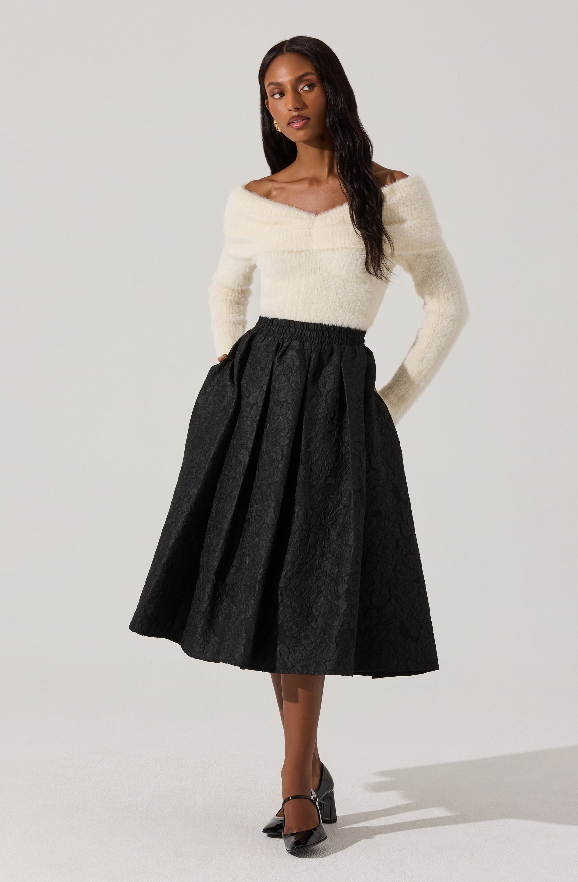 A woman is wearing a white off-the-shoulder, long-sleeve sweater with a fuzzy texture. She pairs it with a high-waisted, black, pleated midi skirt that has a subtle floral pattern. She completes her outfit with black high-heeled shoes featuring ankle straps.