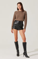 A woman is wearing a brown cable-knit sweater with pearl embellishments on the shoulders, paired with high-waisted black leather shorts. She is also wearing tall black leather heeled boots and gold hoop earrings.