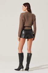 A woman is wearing a taupe-colored, chunky knit sweater adorned with pearl embellishments on the shoulders. She has paired it with high-waisted black leather shorts and knee-high black leather boots with stiletto heels.