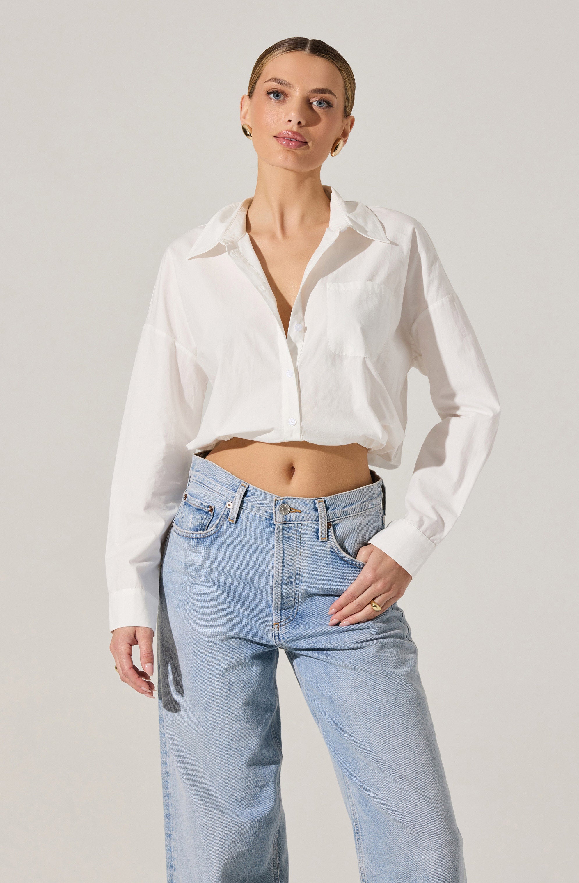 A woman is wearing a white, long-sleeved, button-down shirt that is slightly oversized and tied at the waist, paired with light blue, high-waisted jeans. The shirt has a classic collar and a front pocket on the left side.