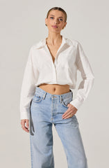 A woman is wearing a white, long-sleeved, button-down shirt that is slightly oversized and tied at the waist, paired with light blue, high-waisted jeans. The shirt has a classic collar and a front pocket on the left side.